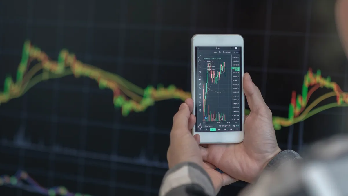 Trader holding a phone with a crypto price chart in front of a trading screen