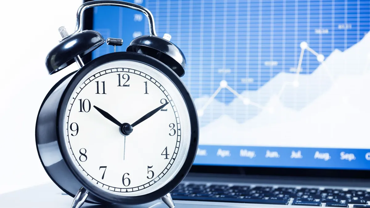 Alarm clock in front of a laptop showing a rising market chart
